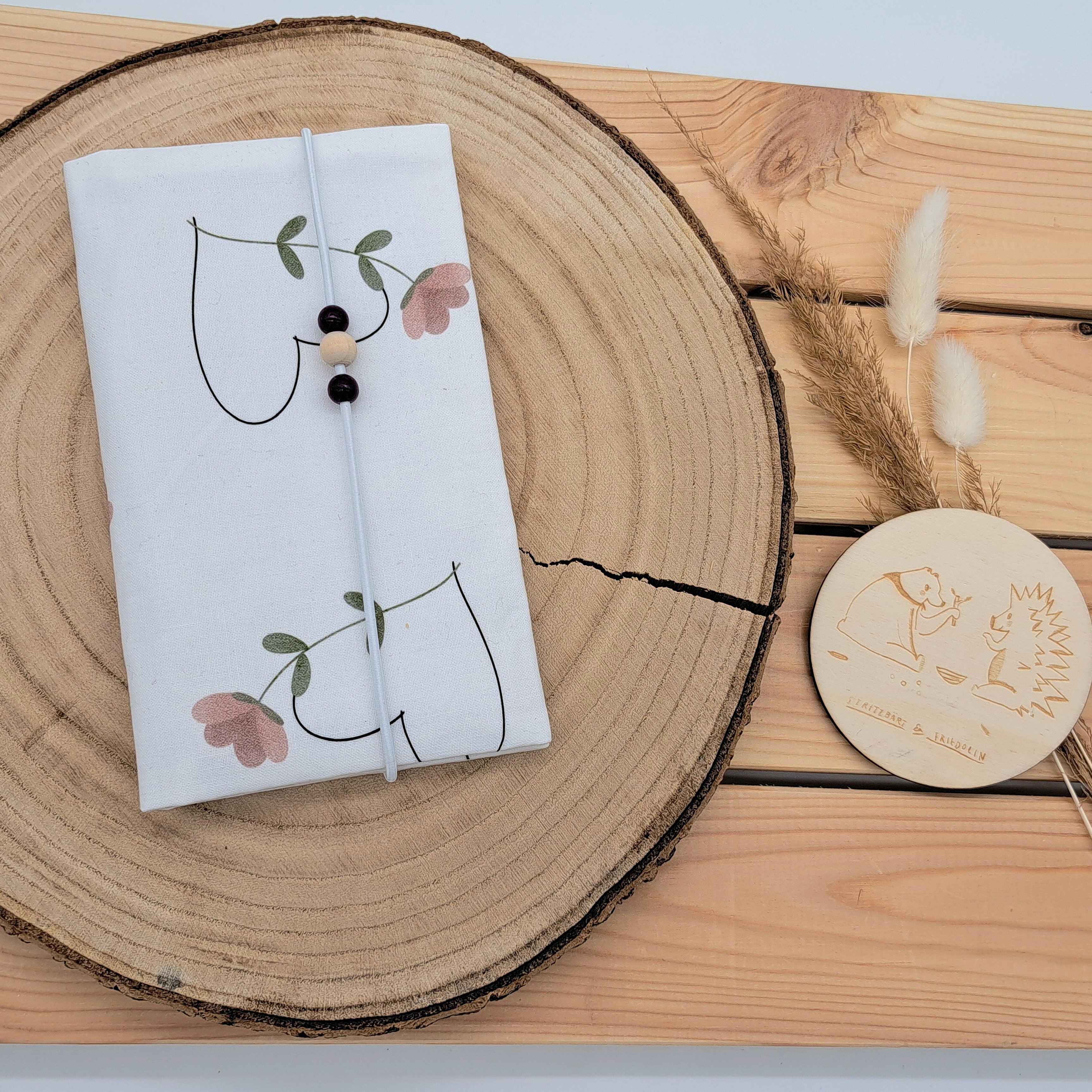 Handgefertigte Mutter-Kind-Pass-Hülle aus biozertifizierter Baumwolle mit E-Card-Fach, Gummiband-Verschluss und dekorativen Holzperlen – nachhaltig, stilvoll und praktisch. Schlichtes Design mit einem Herz aus Blumen in Rosa und Grün.