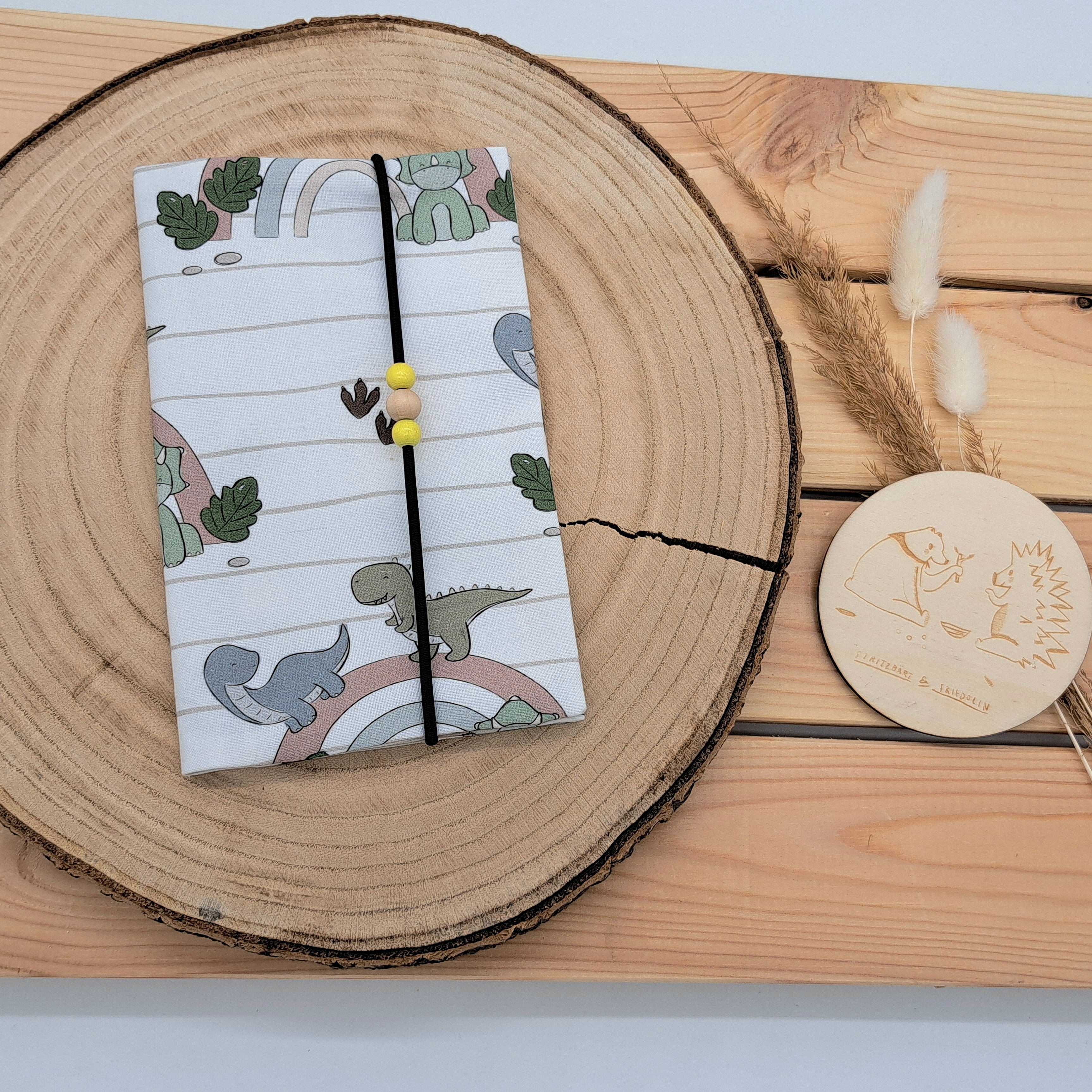 Handgefertigte Mutter-Kind-Pass-Hülle aus biozertifizierter Baumwolle mit E-Card-Fach, Gummiband-Verschluss und dekorativen Holzperlen – nachhaltig, stilvoll und praktisch. Niedliches und neutrales Design mit Dinos die über einen Regenbogen spazieren. Dazu einen schlichten Hintergrund mit Linien und Blätter.