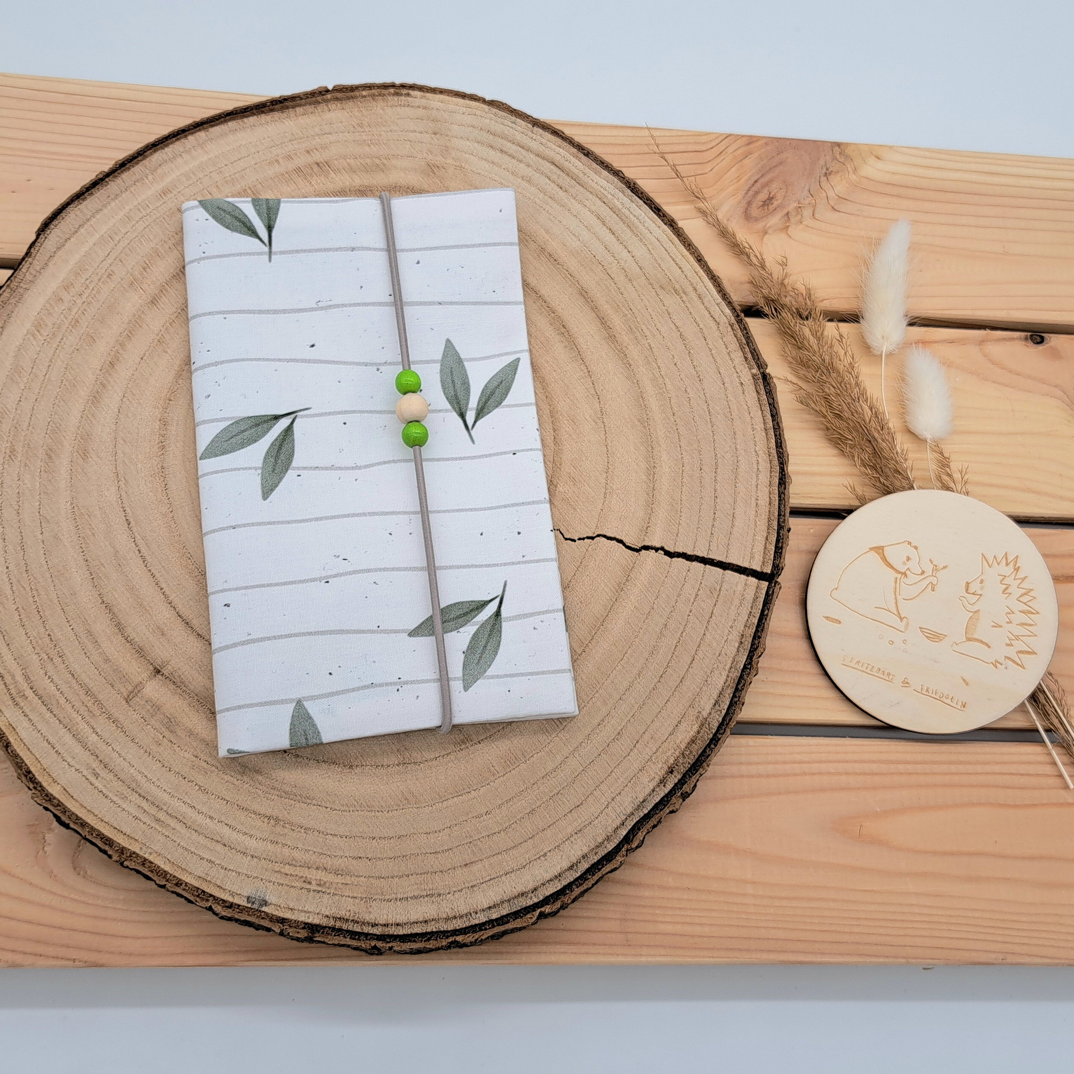 Handgefertigte Mutter-Kind-Pass-Hülle aus biozertifizierter Baumwolle mit E-Card-Fach, Gummiband-Verschluss und dekorativen Holzperlen – nachhaltig, stilvoll und praktisch. Schlichtes und neutrales Design mit weißem Hintergrund und leicht gräulichen Linien. Darauf zu sehen sind grüne Blätter.