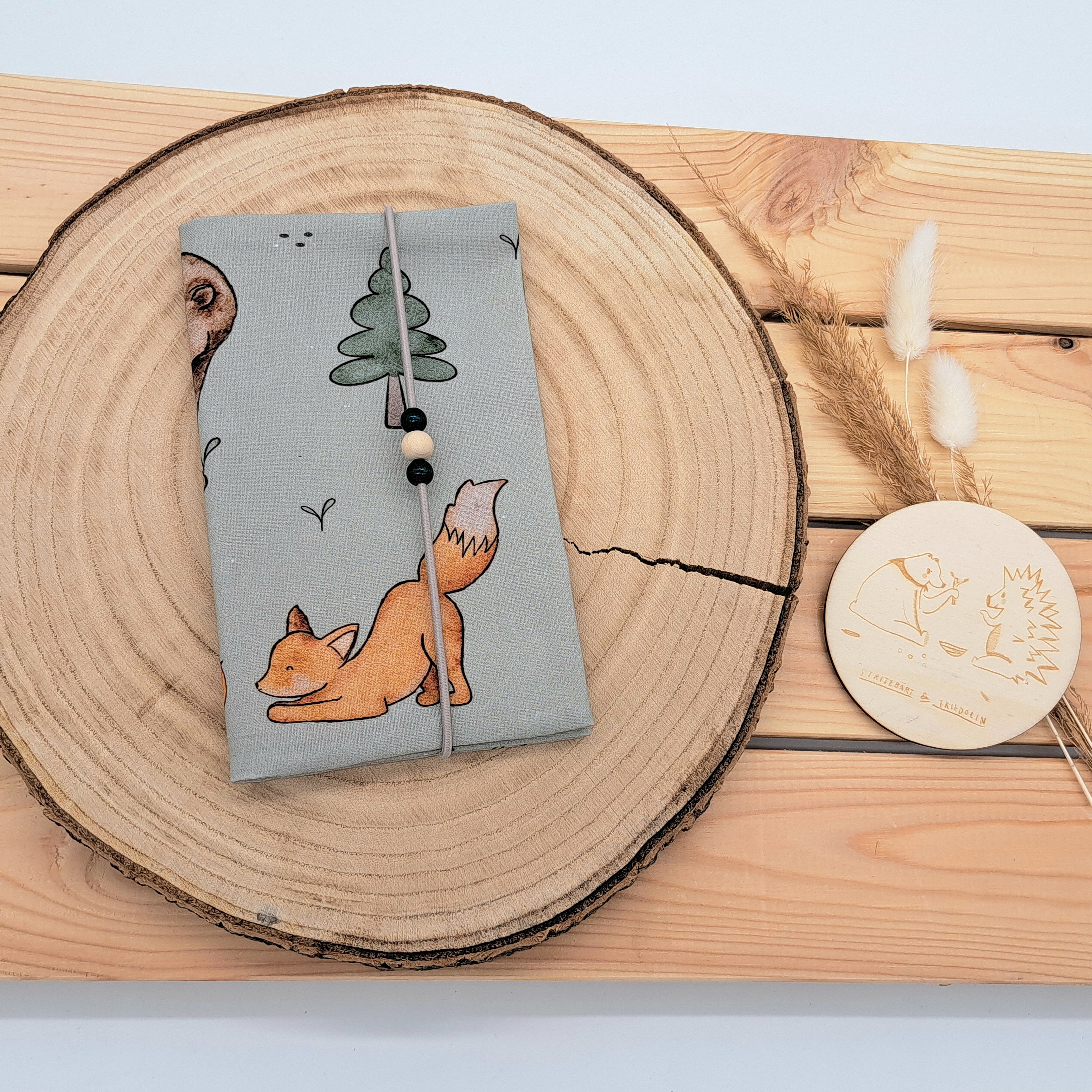 Handgefertigte Mutter-Kind-Pass-Hülle aus biozertifizierter Baumwolle mit E-Card-Fach, Gummiband-Verschluss und dekorativen Holzperlen – nachhaltig, stilvoll und praktisch. Neutraler Hintergrund in olivgrün. Darauf sind Bäume, ein Fuchs und ein Bär zu sehen.