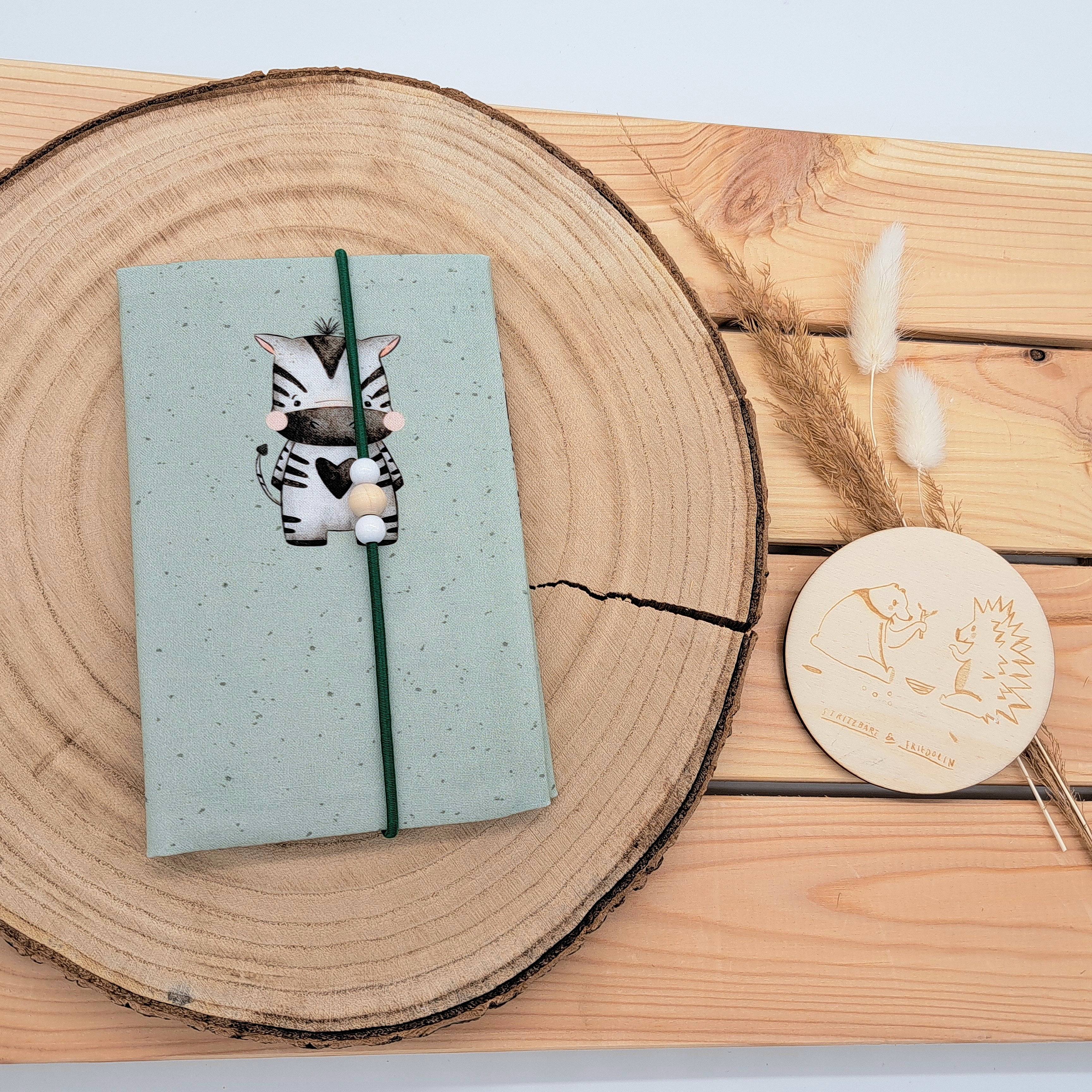Handgefertigte Mutter-Kind-Pass-Hülle aus biozertifizierter Baumwolle mit E-Card-Fach, Gummiband-Verschluss und dekorativen Holzperlen – nachhaltig, stilvoll und praktisch. Neutrales Motiv in zarten Grün gehalten. Darauf ein kleines süßes Zebra.