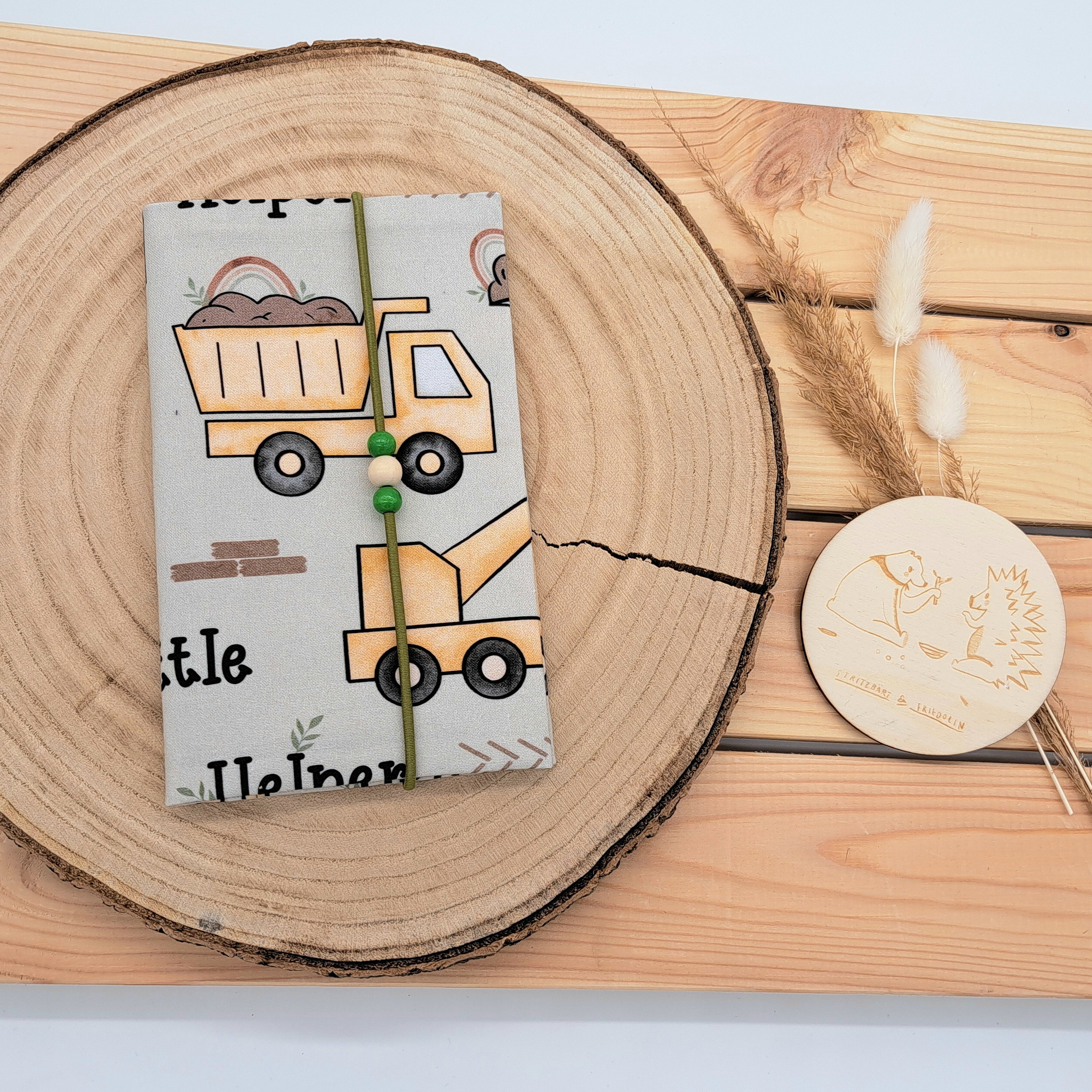 Handgefertigte Mutter-Kind-Pass-Hülle aus biozertifizierter Baumwolle mit E-Card-Fach, Gummiband-Verschluss und dekorativen Holzperlen – nachhaltig, stilvoll und praktisch. Schlichtes Design mit ockerfarbenen Baustellenfahrzeugen und der Aufschrift 