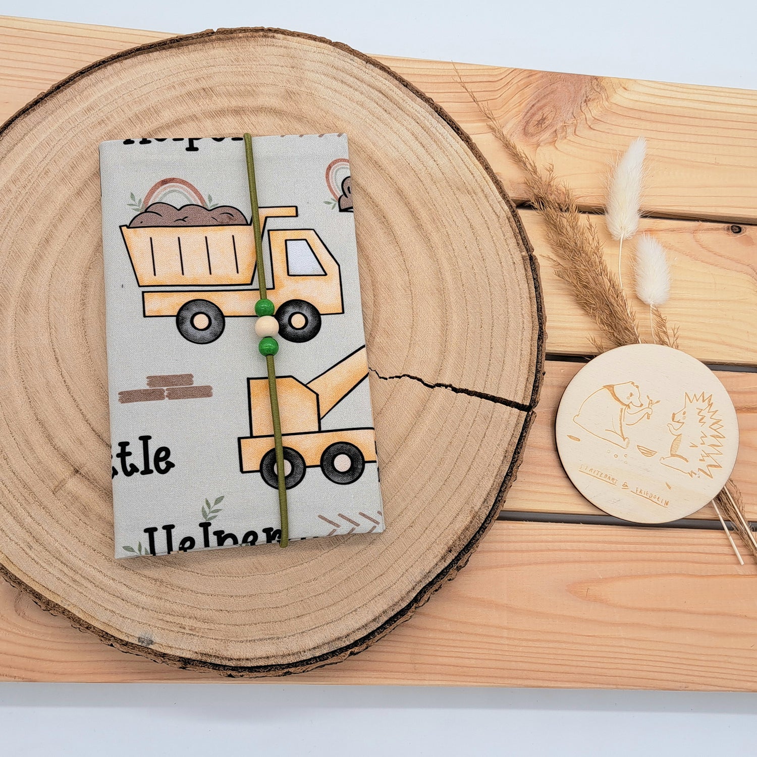 Handgefertigte Mutter-Kind-Pass-Hülle aus biozertifizierter Baumwolle mit E-Card-Fach, Gummiband-Verschluss und dekorativen Holzperlen – nachhaltig, stilvoll und praktisch. Schlichtes Design mit ockerfarbenen Baustellenfahrzeugen und der Aufschrift "little Helper".