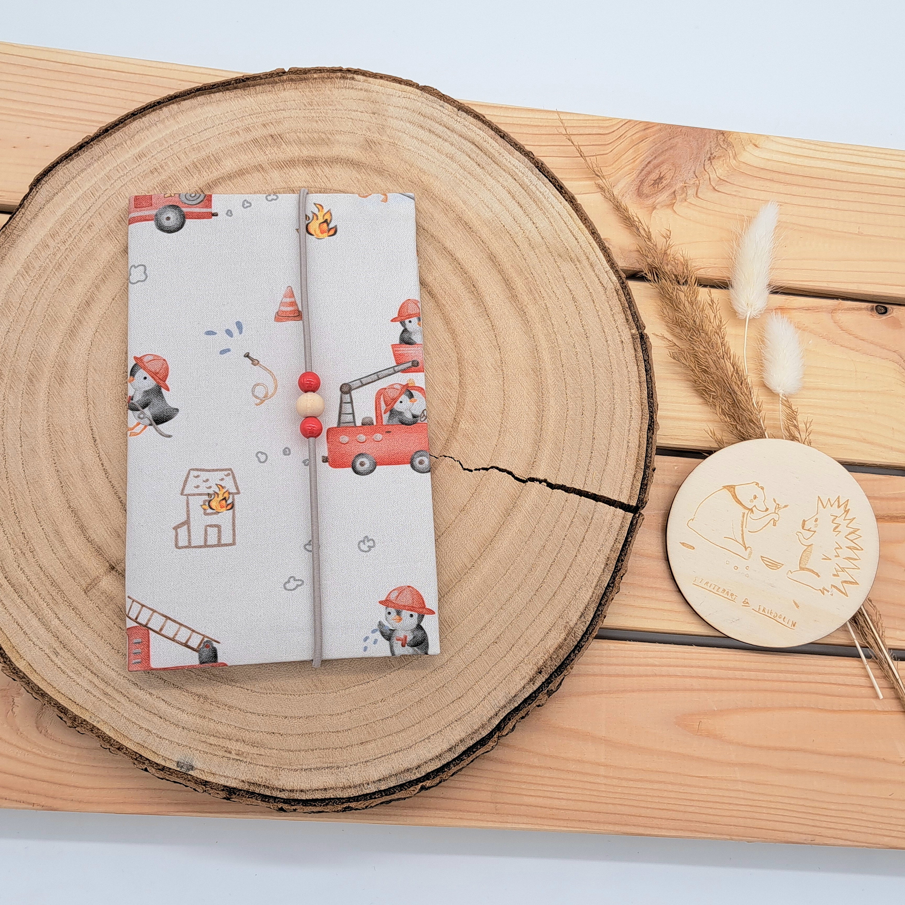 Handgefertigte Mutter-Kind-Pass-Hülle aus biozertifizierter Baumwolle mit E-Card-Fach, Gummiband-Verschluss und dekorativen Holzperlen – nachhaltig, stilvoll und praktisch. Süßes Feuerwehrdesign mit Pinguinen, Feuerwehrautos, Schlauch, Feuer und Wasser.
