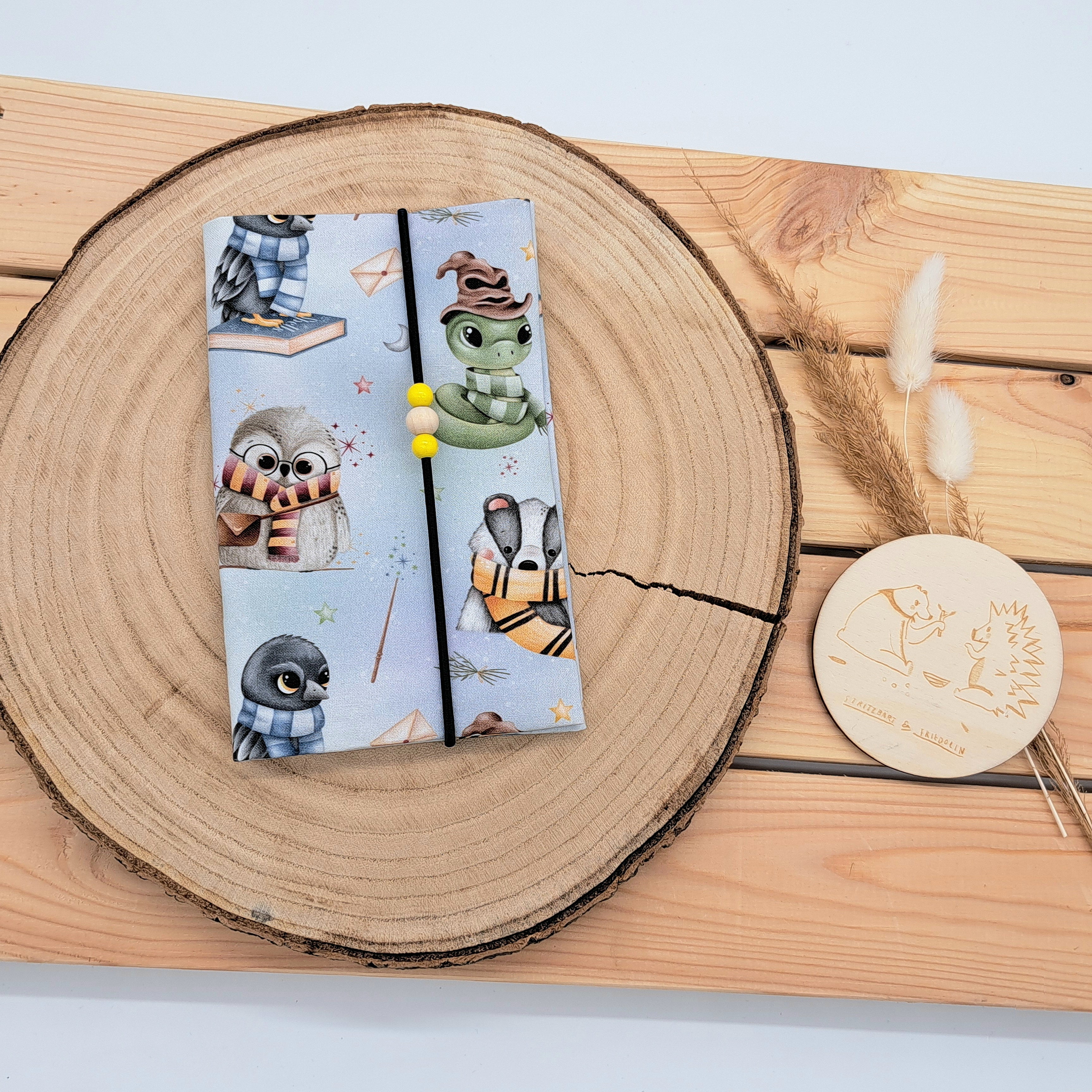 Handgefertigte Mutter-Kind-Pass-Hülle aus biozertifizierter Baumwolle mit E-Card-Fach, Gummiband-Verschluss und dekorativen Holzperlen – nachhaltig, stilvoll und praktisch. Motiv im Zauberstil. Blauer Hintergrund mit Briefkuvert, Zauberstab und Funkelsternen. Darauf sind verschiedene Tiere im Zauberstil. (Eule, Rabe, Schlange, Dachs)
