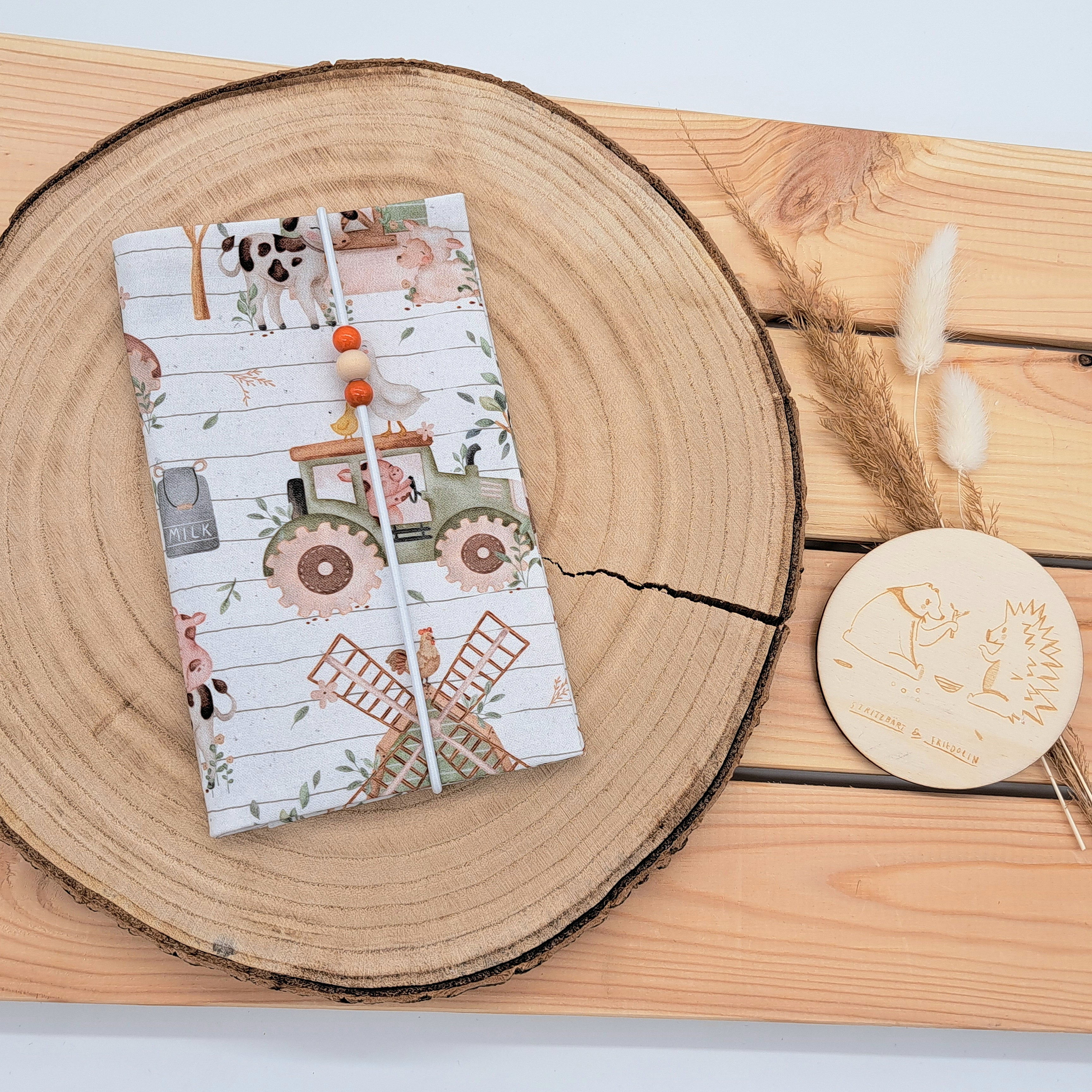 Handgefertigte Mutter-Kind-Pass-Hülle aus biozertifizierter Baumwolle mit E-Card-Fach, Gummiband-Verschluss und dekorativen Holzperlen – nachhaltig, stilvoll und praktisch. Neutrales Bauernhofmuster mit Traktor, Gans, Schwein, Kuh, Mühle.