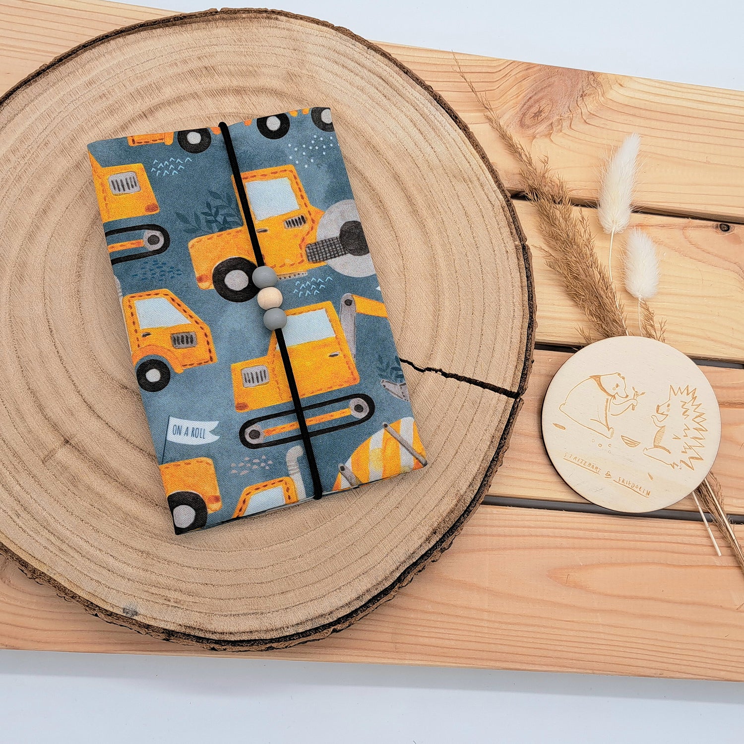 Handgefertigte Mutter-Kind-Pass-Hülle aus biozertifizierter Baumwolle mit E-Card-Fach, Gummiband-Verschluss und dekorativen Holzperlen – nachhaltig, stilvoll und praktisch. EINZELSTÜCK  mit Baustellenfahrzeugen.
