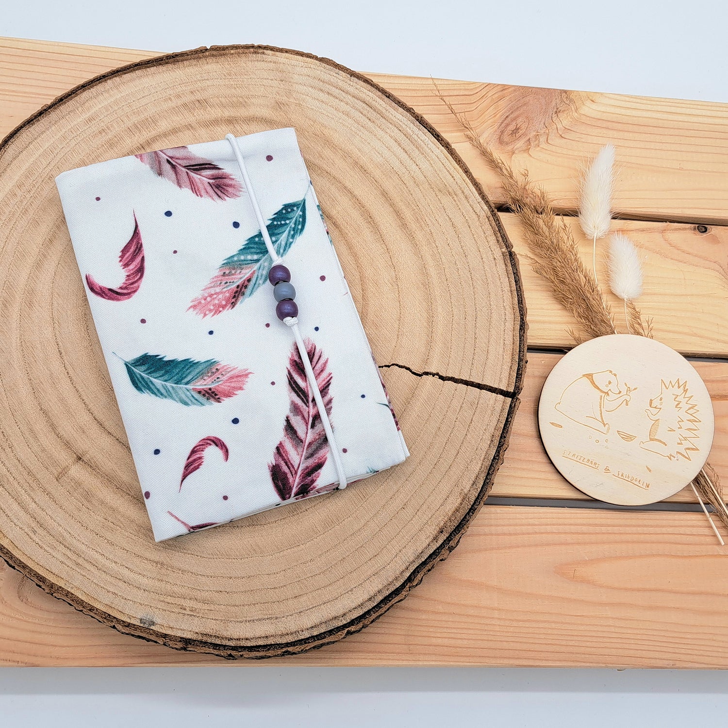 Handgefertigte Mutter-Kind-Pass-Hülle aus biozertifizierter Baumwolle mit E-Card-Fach, Gummiband-Verschluss und dekorativen Holzperlen – nachhaltig, stilvoll und praktisch. EINZELSTÜCK mit lilafarbenen Federn.