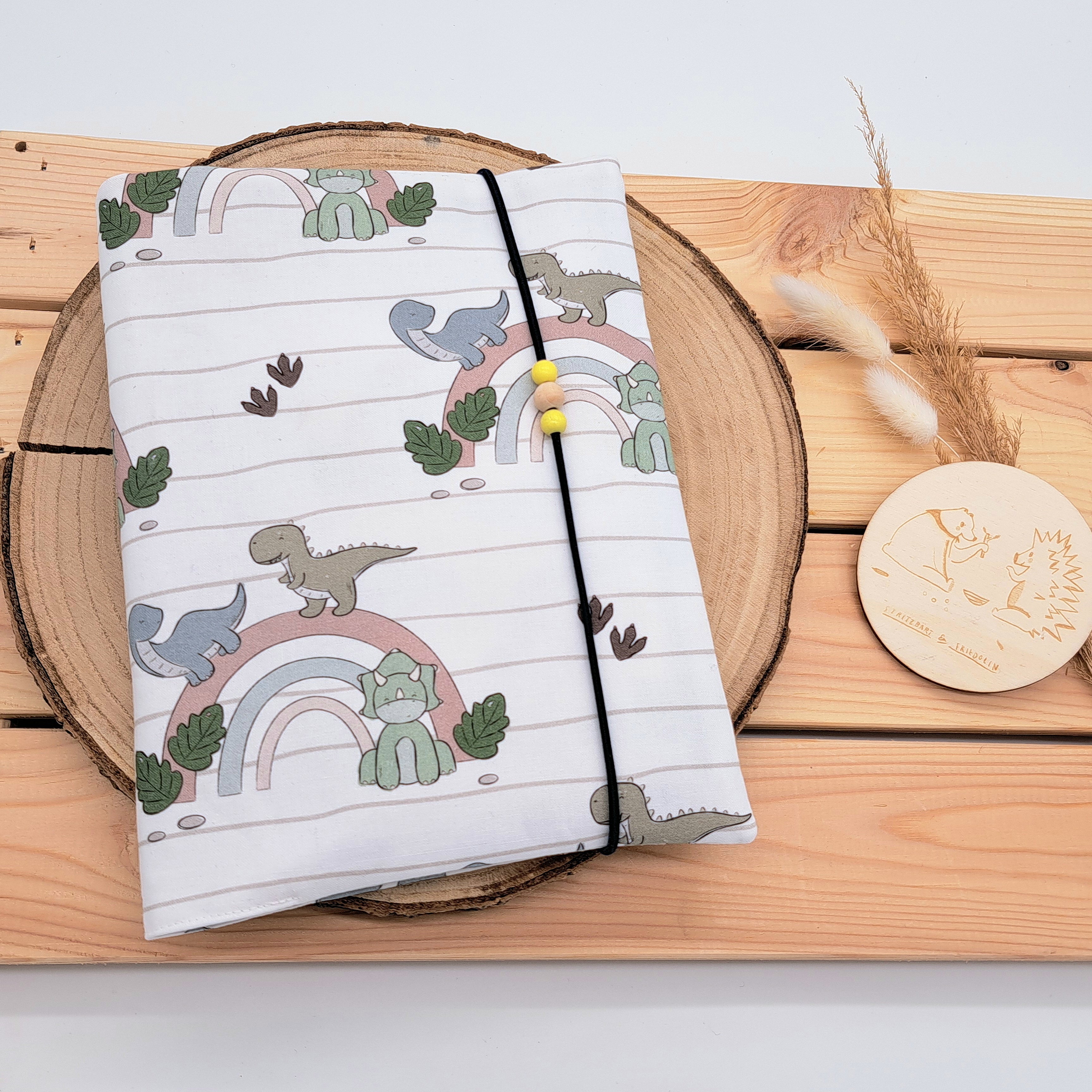 Handgefertigte Windeltasche aus 100% Bio-Baumwolle mit Fach für Windeln und Feuchttücher, Gummikordel-Verschluss mit Holzperlen; nachhaltiges und praktisches Accessoire für Eltern unterwegs. Niedliches Dino-Design, die über einen schlichten Regenbogen spazieren.