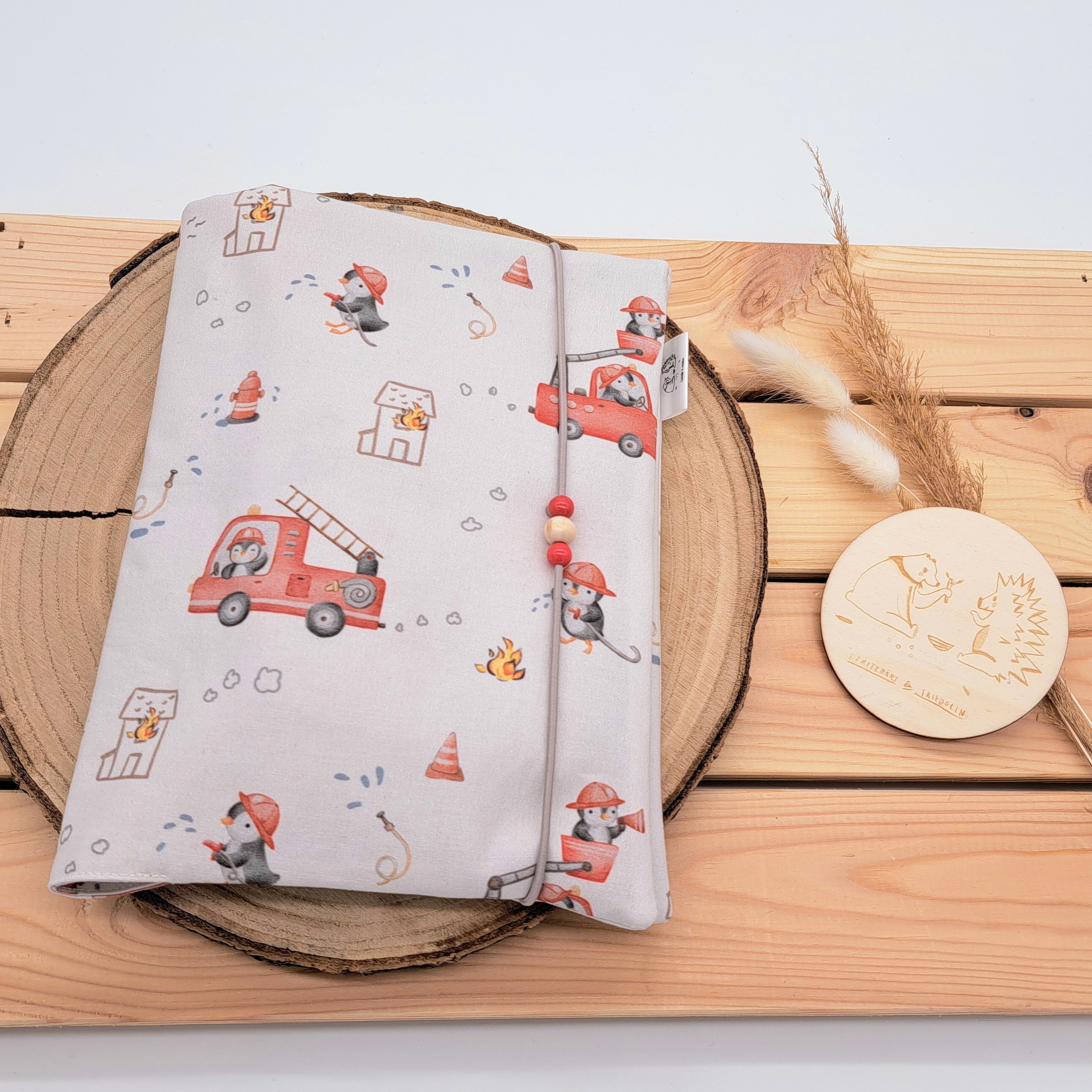 Handgefertigte Windeltasche aus 100% Bio-Baumwolle mit Fach für Windeln und Feuchttücher, Gummikordel-Verschluss mit Holzperlen; nachhaltiges und praktisches Accessoire für Eltern unterwegs. Neutrales und süßes Design im Feuerwehrstil.Kleine Pinguine verkleidet als Feuerwehrhelden mit Autos, Wasserschlauch und kleinem Feuer.