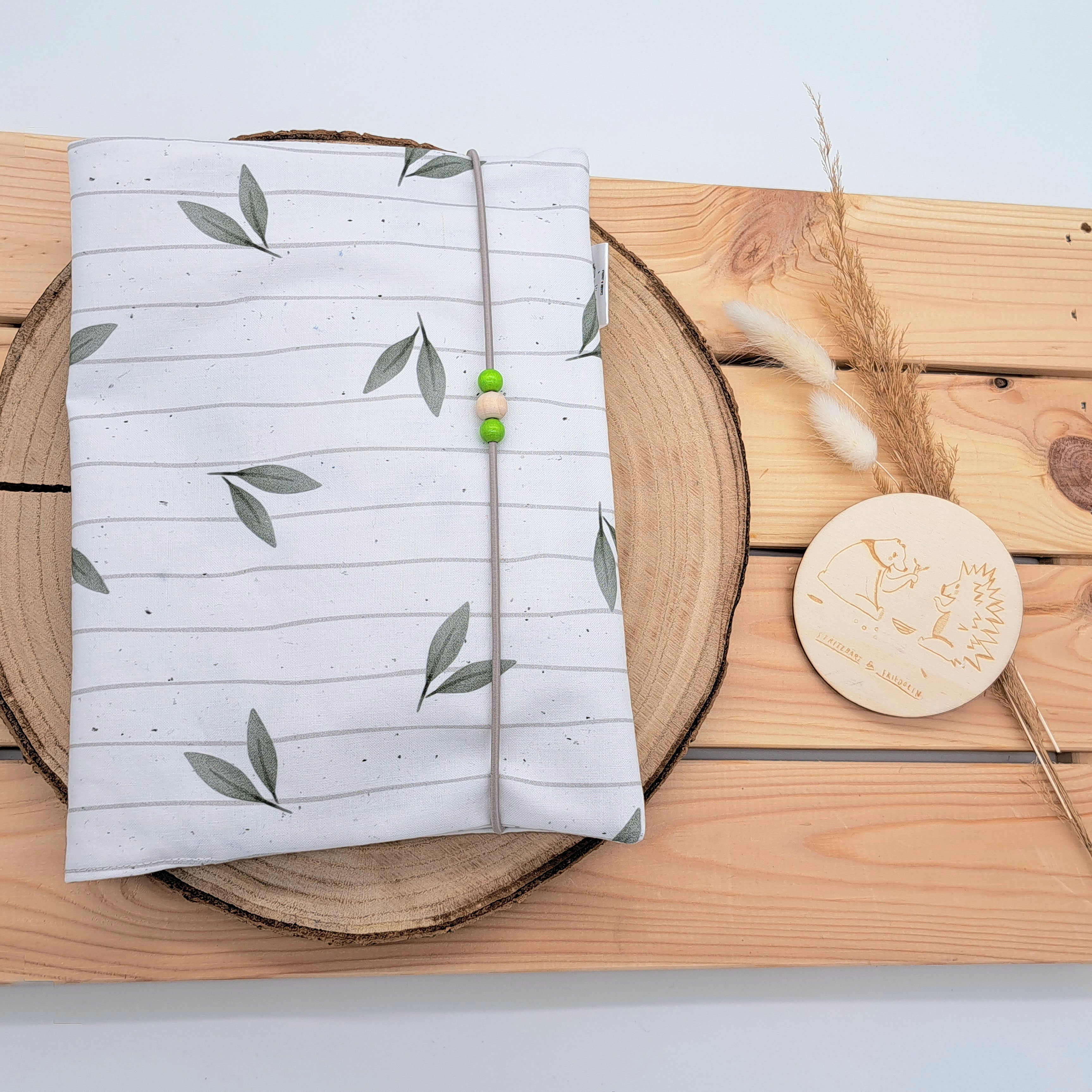 Handgefertigte Windeltasche aus 100% Bio-Baumwolle mit Fach für Windeln und Feuchttücher, Gummikordel-Verschluss mit Holzperlen; nachhaltiges und praktisches Accessoire für Eltern unterwegs. Neutrales und schlichtes Design auf weiß gestreiftem Hintergrund. Darauf grüne Blätter.