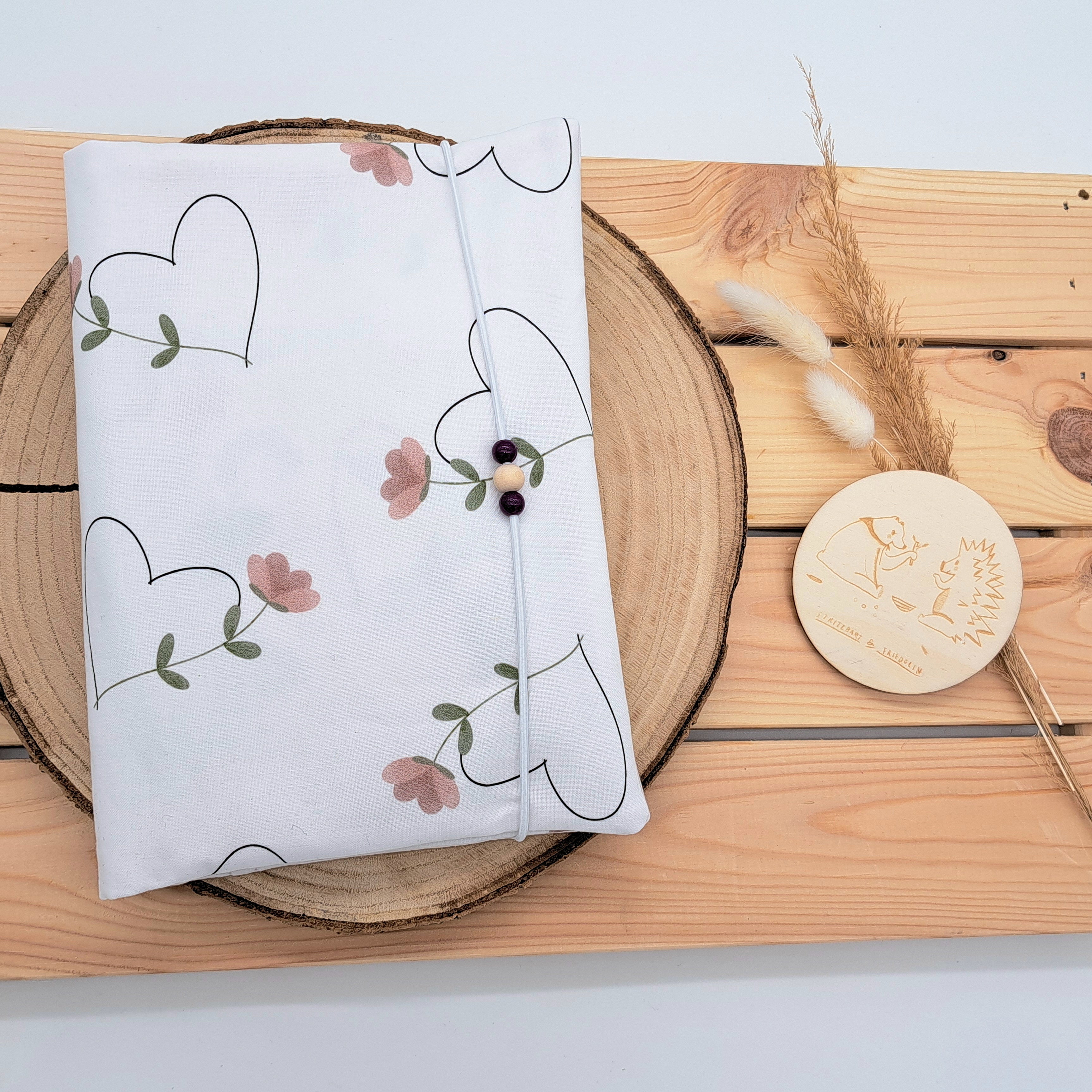 Handgefertigte Windeltasche aus 100% Bio-Baumwolle mit Fach für Windeln und Feuchttücher, Gummikordel-Verschluss mit Holzperlen; nachhaltiges und praktisches Accessoire für Eltern unterwegs. Schlichtes Design mit Herzen aus zarten rosa Blumen.