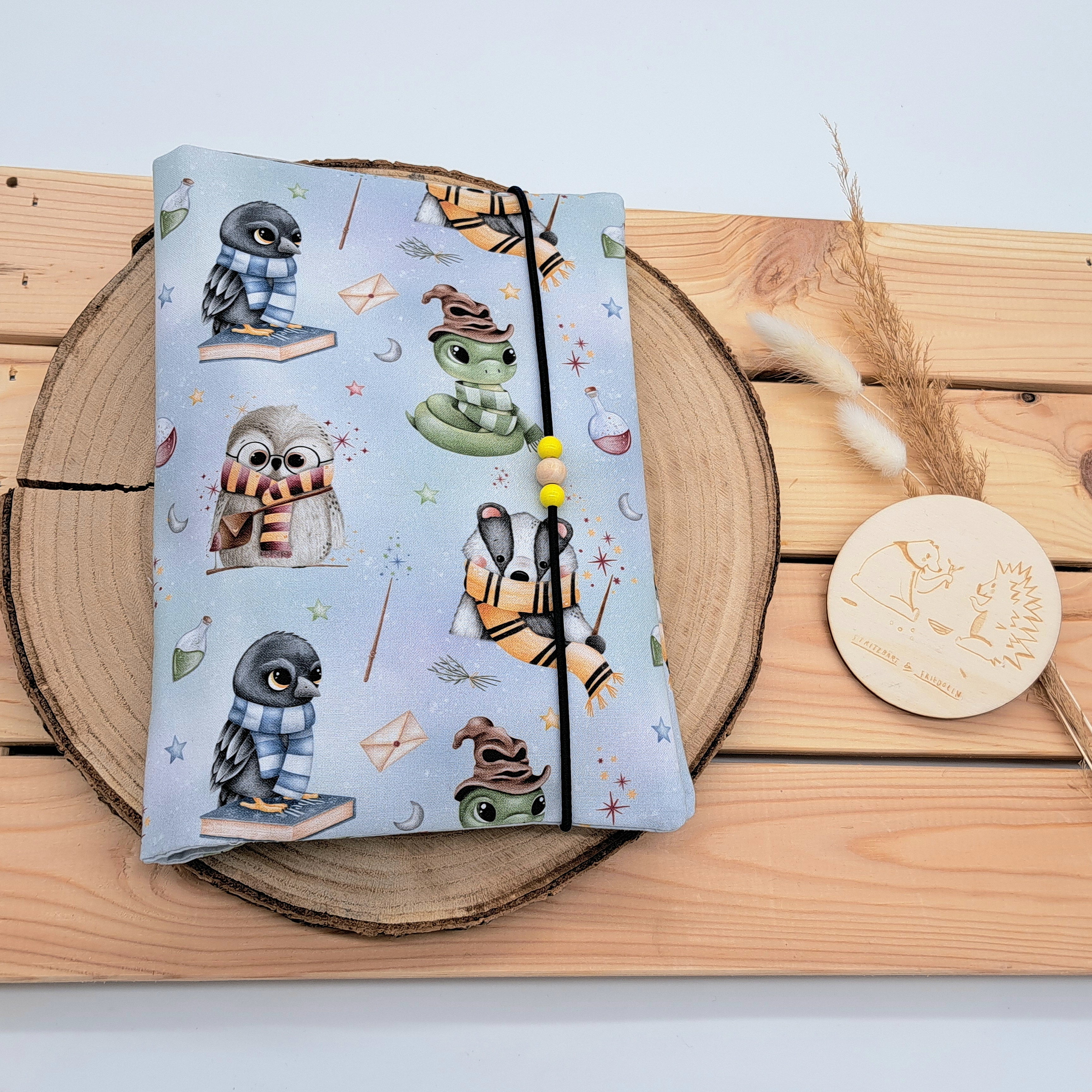 Handgefertigte Windeltasche aus 100% Bio-Baumwolle mit Fach für Windeln und Feuchttücher, Gummikordel-Verschluss mit Holzperlen; nachhaltiges und praktisches Accessoire für Eltern unterwegs. Zaubertiere mit Zauberstäbe, Briefumschläge und Zaubertränke auf blauverwischten Hintergrund. Die Tiere sind Dachs, Rabe, Eule und Schlange. 