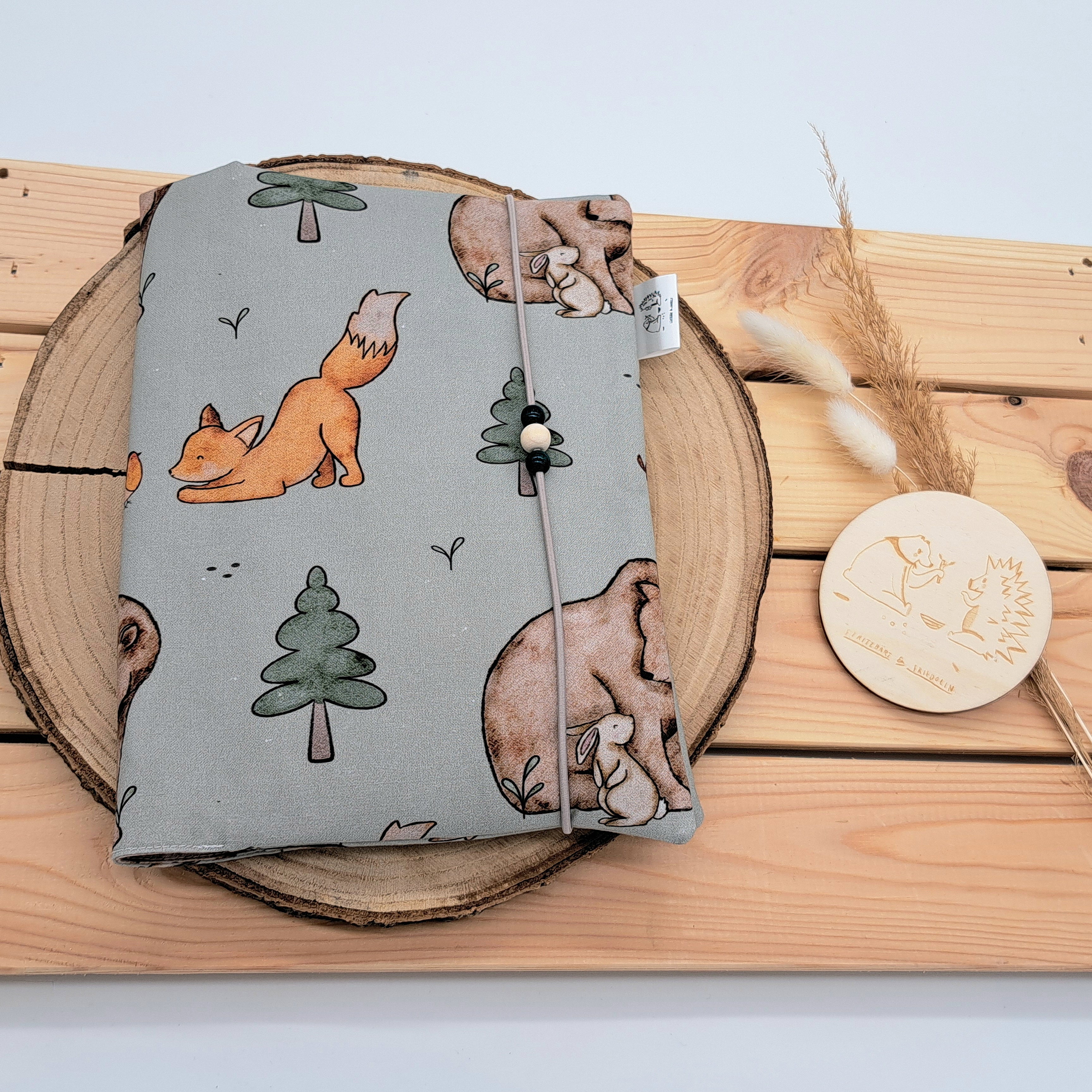 Handgefertigte Windeltasche aus 100% Bio-Baumwolle mit Fach für Windeln und Feuchttücher, Gummikordel-Verschluss mit Holzperlen; nachhaltiges und praktisches Accessoire für Eltern unterwegs. Auf einem grünen Hintergrund spielen hier ein Fuchs, Bär und Hase inmitten von Bäumen.