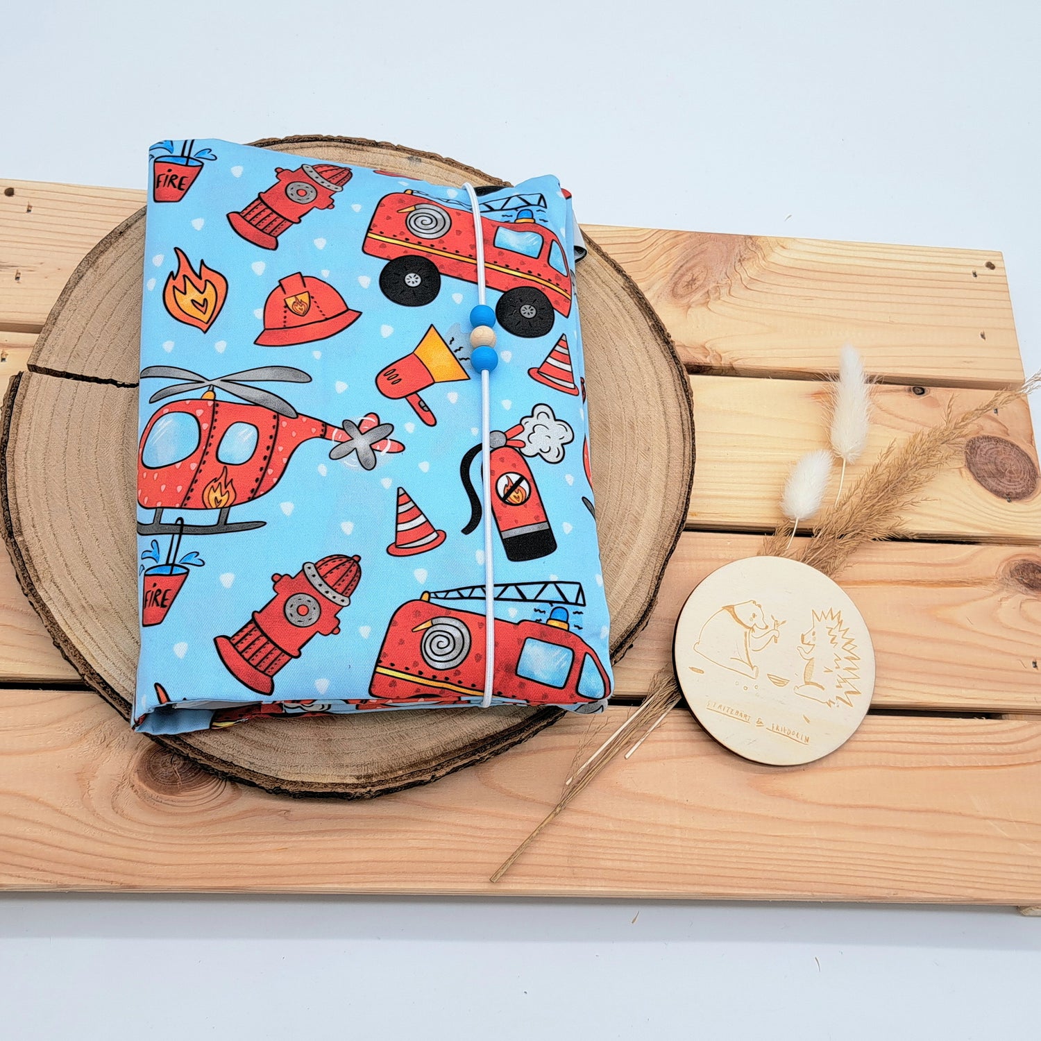 Handgefertigte Windeltasche aus 100% Bio-Baumwolle mit Fach für Windeln und Feuchttücher, Gummikordel-Verschluss mit Holzperlen; nachhaltiges und praktisches Accessoire für Eltern unterwegs. EINZELSTÜCK. Auf blauen Hintergrund sind rote Feuerwehrautos, Helme, Hydranten und ein roter Feuerwehrhubschrauber zu sehen.
